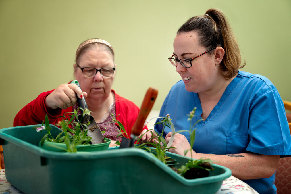 Two women gardening together, handling tools and plant cuttings over a green planter tray.