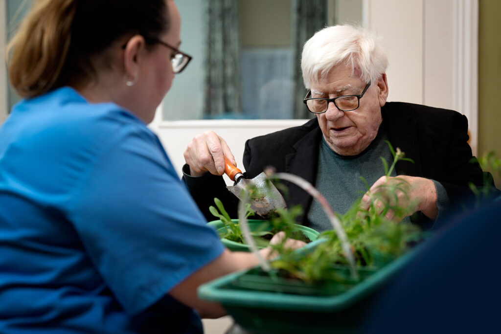 Elderly man with white hair and glasses plants herbs, aided by a caregiver with a small trowel over a green planter.