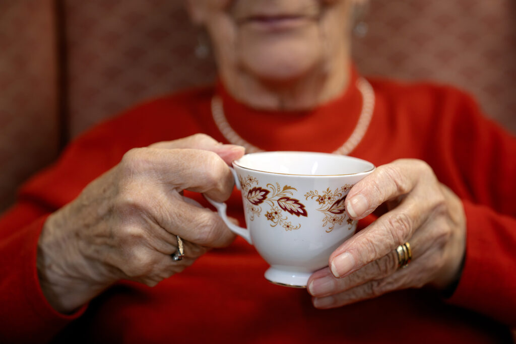 An elderly person in a red sweater holds a white teacup with gold floral pattern, wearing a pearl necklace.