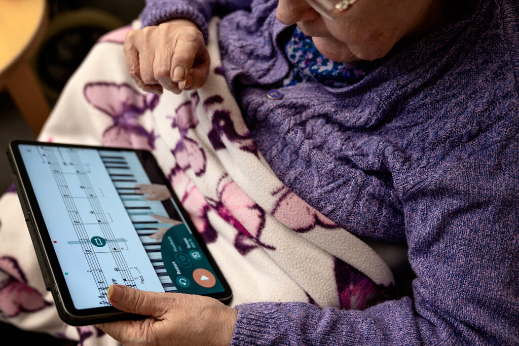 Older person in a purple sweater uses a tablet to view piano sheet music on a floral blanket area behind them.