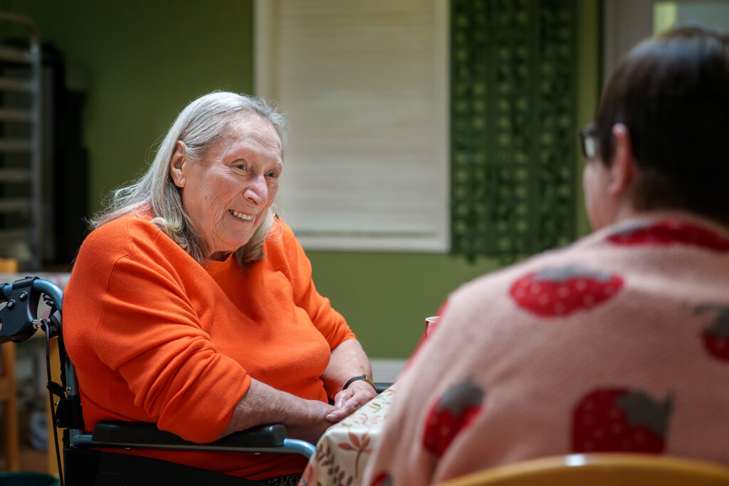 Smiling elderly woman in an orange top sits in a wheelchair at a table, chatting with another person indoors.